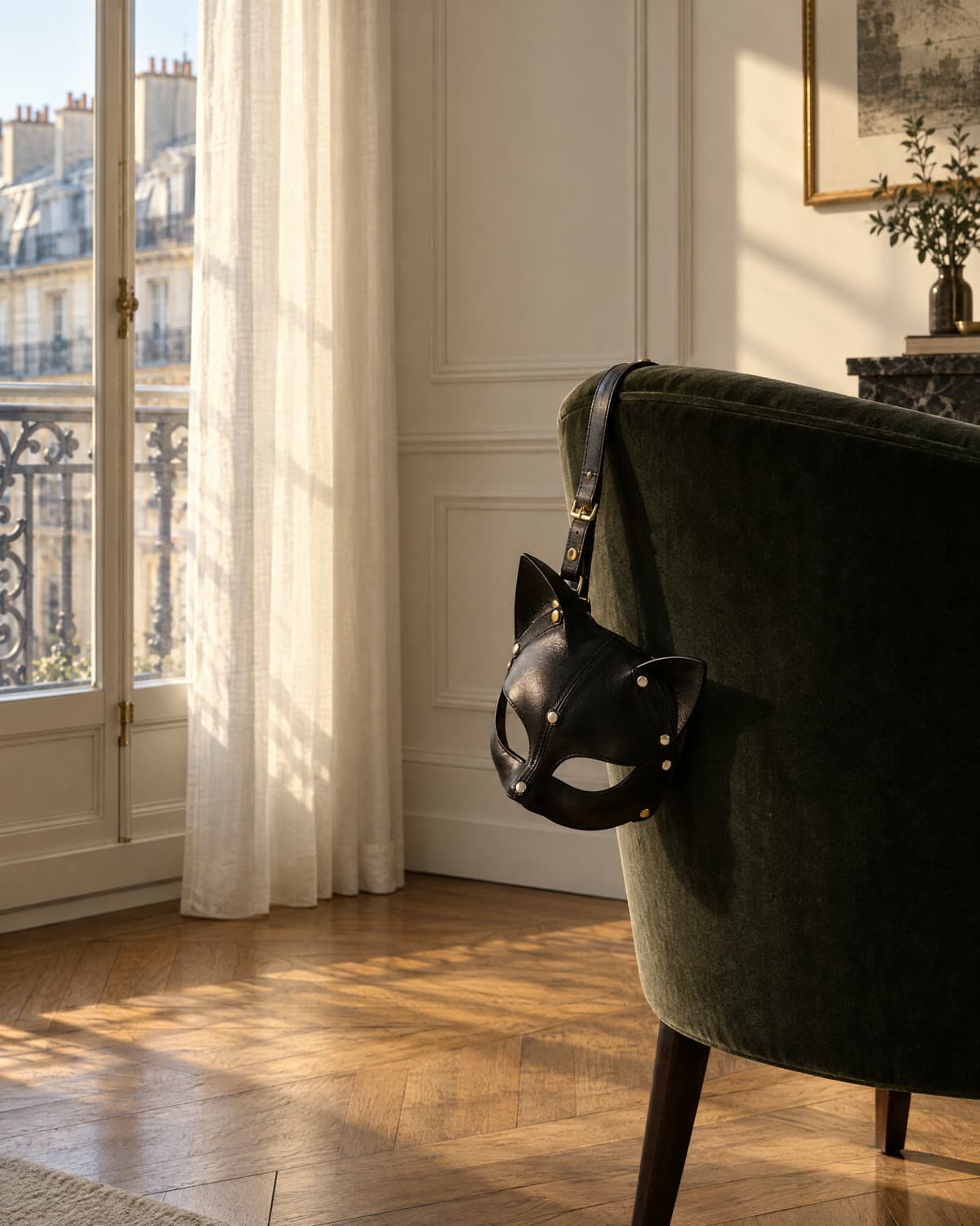 Masque de chat en cuir noir suspendu au dossier d'un fauteuil de velours, devant une fenêtre parisienne